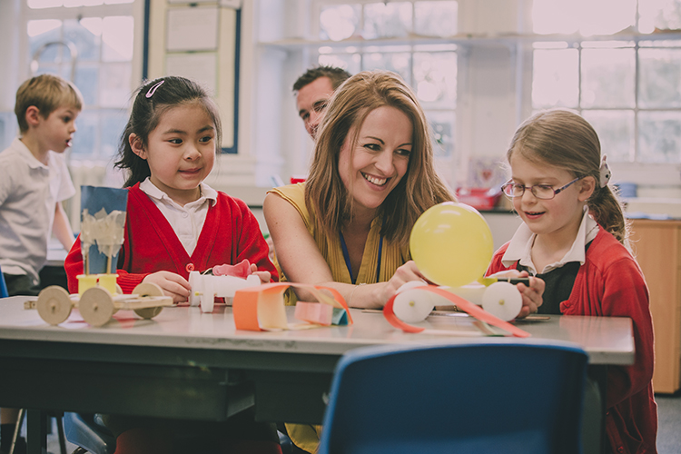 Primary school teacher is helping two of her students with a STEM project. They are building something using recycled items and crafts equipment.